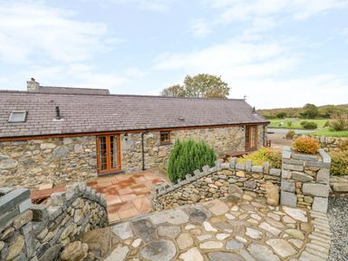 An exterior view of a stone cottage with a tiled roof surrounded by a stone patio and steps with greenery in the background at Poppy Cottage in Caeathro near Caernarfon