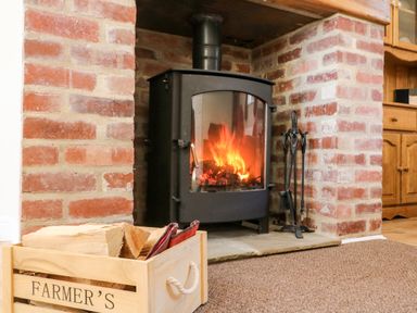 A wood burning stove with fire inside a brick fireplace with fire tools and a wooden box of firewood at 2 Pines Farm Cottages in Ulleskelf