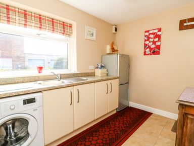 A kitchen with a washing machine, sink, refrigerator, countertop, red patterned rug, window with a plaid blind, and decorations on the walls at 2 Pines Farm Cottages Ulleskelf