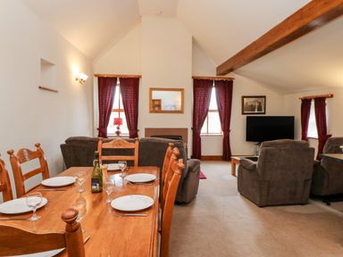 A dining area with a wooden table set with plates and glasses adjacent to a sitting area with sofas a flat screen TV and windows with curtains at The Dairy in Milton near Brampton