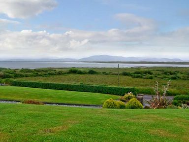 A grassy yard with bushes and a hedge overlooking a body of water and distant hills at Heron's Nest in Inch County Kerry