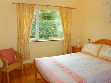 A bedroom with a bed a wooden bedside table with a lamp a wicker chair with a cushion and a window with curtains at Heron's Nest in Inch County Kerry