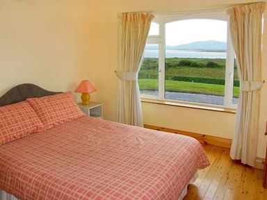 A bedroom with a bed covered in patterned bedding and a window with curtains showing a view of grass and hills at Heron's Nest in Inch County Kerry