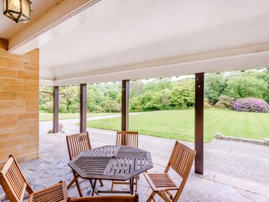 A covered outdoor patio with a wooden table and four chairs overlooking a lawn and trees at Plas Gwynfryn in Llanbedr