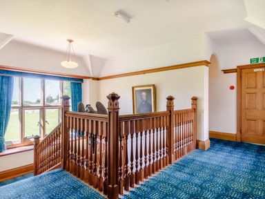 A stairway landing with wooden railing a portrait on the wall blue carpet and a large window with blue curtains at Plas Gwynfryn in Llanbedr