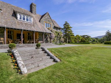 A stone house with wooden chairs on a porch steps leading to a lawn and trees at Plas Gwynfryn in Llanbedr