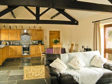 An open plan kitchen and living area with wooden cabinets a dining table with chairs and a black sofa with cushions and a throw at The Byre in Llanddewi Skirrid near Abergavenny