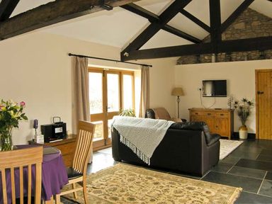 A living room with a dining table near wooden doors and a black sofa under exposed ceiling beams at The Byre in Llanddewi Skirrid near Abergavenny