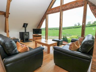 A living room with black leather sofas wooden coffee table wood stove and large windows overlooking a green field at Shepherd's Barn Between Milborne St Andrew and Winterborne Whitechurch
