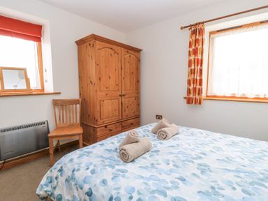 A bedroom with a bed with towels on it a wooden wardrobe a wooden chair and two windows with curtains at Shepherd's Barn Between Milborne St Andrew and Winterborne Whitechurch