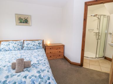 A bedroom with a bed with blue patterned bedding a wooden bedside table with a lamp and an adjoining bathroom with a glass shower at Shepherd's Barn Between Milborne St Andrew and Winterborne Whitechurch