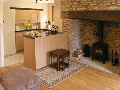 A kitchen area with wooden cabinets and a kettle beside a stone fireplace with a wood stove and firewood at Candy Cottage Thornford near Sherborne