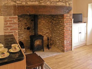 A room with a wood-burning stove set in a brick and stone fireplace a wooden table cups on a countertop and a television on a white cabinet at Candy Cottage Thornford near Sherborne