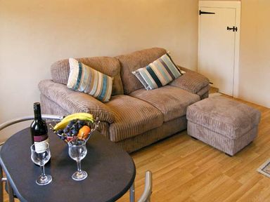 A living room with a brown sofa, striped cushions, ottoman, round table with wine bottle, glasses and fruit bowl at Candy Cottage Thornford near Sherborne