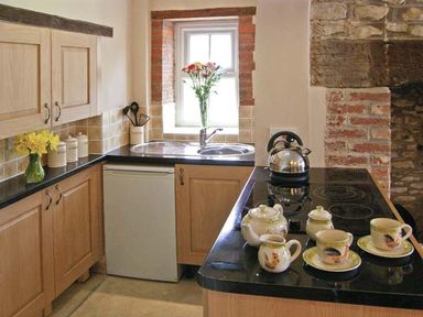 A kitchen with wooden cabinets a black countertop a sink a window with flowers a kettle and a tea set on the counter at Candy Cottage Thornford near Sherborne