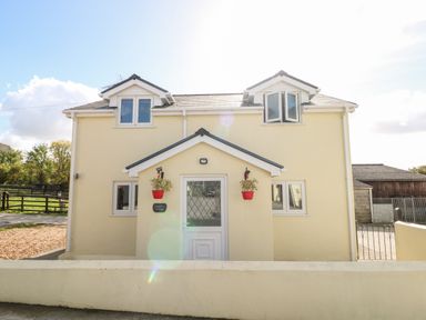 A two-story pale yellow house with white framed windows and a white front door with two red flower pots on either side of the entrance at Saddlers Cottage Berllandeg Farm