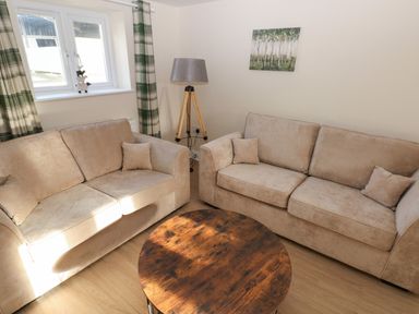 A living room with two beige sofas a round wooden coffee table a tripod floor lamp a painting of trees on the wall and a window with green plaid curtains at Saddlers Cottage Berllandeg Farm Clunderwen near Narberth