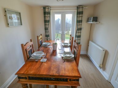 A dining room with a wooden table set with plates glasses and cutlery four chairs and glass doors with plaid curtains at Saddlers Cottage Berllandeg Farm Clunderwen near Narberth