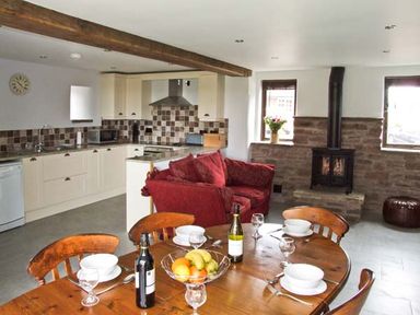 A kitchen with cream cabinets next to a dining table set with glasses bottles and bowls and a red sofa near a stone fireplace at Bushmills in Kerrys Gate near Abbey Dore