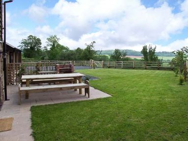 A garden with a wooden picnic table and benches on a patio near a wooden fence and green lawn at Bushmills in Kerry's Gate near Abbey Dore