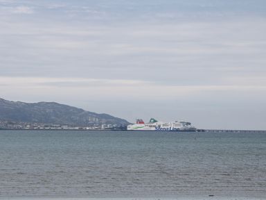 A ferry boat on the sea near a mountainous coastline with industrial buildings and a pier