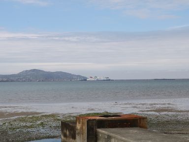 A view of a bay with a ferry in the distance and a rocky shoreline with a concrete structure in the foreground at Ty Fferm Bodlasan in Llanfachraeth near Valley