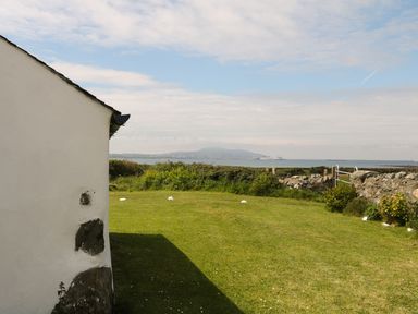 A garden with grass and shrubs overlooking a body of water with a mountain in the distance at Ty Fferm Bodlasan in Llanfachraeth near Valley