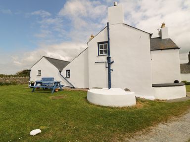 A white building with blue window frames and a blue picnic table on grass at Ty Fferm Bodlasan in Llanfachraeth near Valley