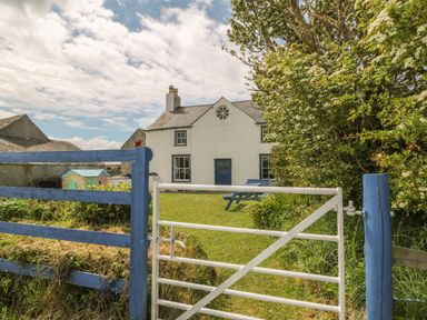 A white house with blue door and window frames behind a white gate and blue fence in a grassy yard at Ty Fferm Bodlasan in Llanfachraeth near Valley