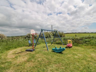 An outdoor play area with a swing set, basketball hoop, two chairs, a small green rocker, and a red toy car on grass at Ty Fferm Bodlasan in Llanfachraeth near Valley