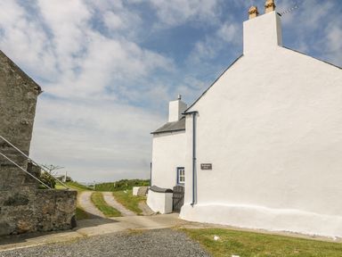 A white building with chimneys next to a gravel and grass driveway under a partly cloudy sky at Ty Fferm Bodlasan in Llanfachraeth near Valley