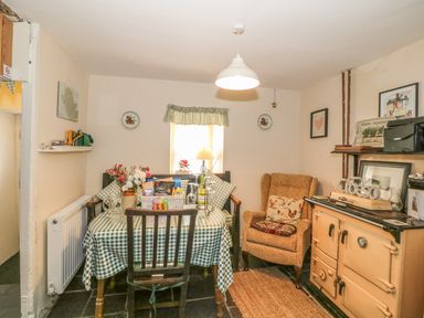 A dining area with a checkered tablecloth on a table flowers drinks a cushioned armchair and an old stove at Ty Fferm Bodlasan in Llanfachraeth near Valley