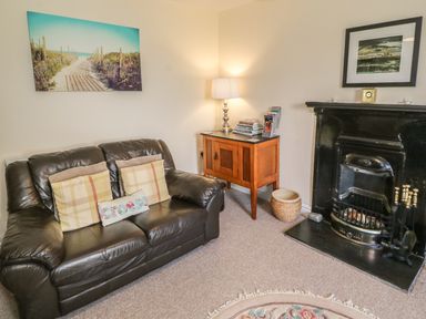 A living room with a leather sofa cushions a wooden side table with a lamp a black fireplace and framed pictures on the wall at Ty Fferm Bodlasan in Llanfachraeth near Valley