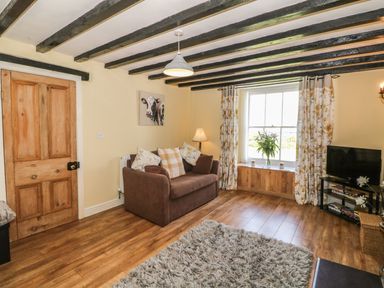 A living room with a brown sofa and cushions a wooden door a window with floral curtains a television on a stand and a rug on the floor at Ty Fferm Bodlasan in Llanfachraeth near Valley