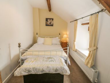 A bedroom with a double bed a small wooden nightstand with lamp yellow check curtains and a flower painting at Ty Fferm Bodlasan in Llanfachraeth near Valley