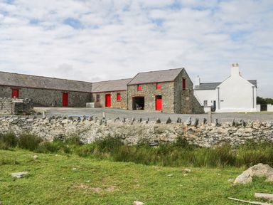 Stone buildings with red doors and windows next to a white house behind a stone wall at Ty Fferm Bodlasan in Llanfachraeth near Valley