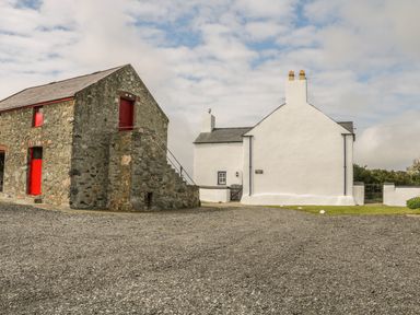 A courtyard with a stone building with red doors and a white house with chimneys at Ty Fferm Bodlasan in Llanfachraeth near Valley