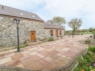 A stone building with wooden doors and windows on a paved courtyard with lamp posts and flower beds at Daffodil Cottage in Caeathro near Caernarfon