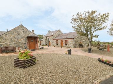 A stone cottage with wooden doors and windows surrounded by gravel and paved walkways with planters and trees at Daffodil Cottage in Caeathro near Caernarfon