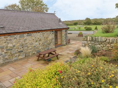 A stone cottage with a tiled roof and wooden picnic table on a paved patio surrounded by plants and a stone wall at Daffodil Cottage in Caeathro near Caernarfon