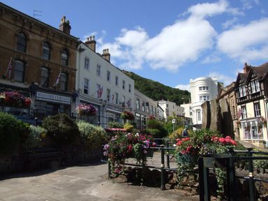 A street scene with flower beds and benches in front of a row of shops and buildings with flags above them in a town