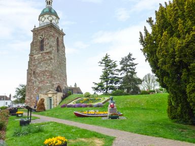 A stone clock tower with a green copper roof in a park with flower beds trees and a bust statue