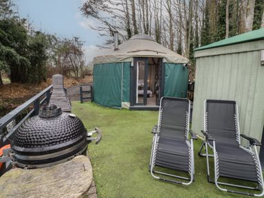 An outdoor area with two black lounge chairs a round yurt with green covering a black barbecue grill and a wooden shed at The Lakeside Yurt in Beckford