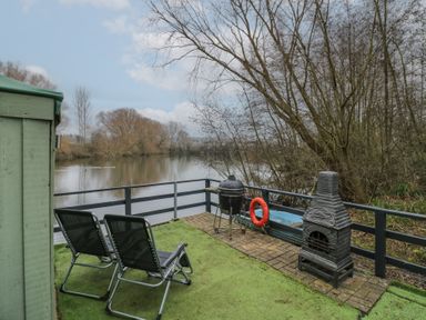 A deck with two lounge chairs a barbecue and a chiminea overlooking a river with trees at The Lakeside Yurt in Beckford