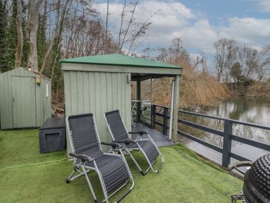 A lakeside deck with two black lounge chairs and a green shed surrounded by trees at The Lakeside Yurt in Beckford