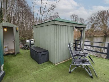 An outdoor area with a small green shed a black storage box two folding chairs a small bathroom with a toilet and a river in the background at The Lakeside Yurt in Beckford