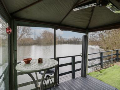A covered deck with a round table and stool overlooking a lake with leafless trees at The Lakeside Yurt in Beckford