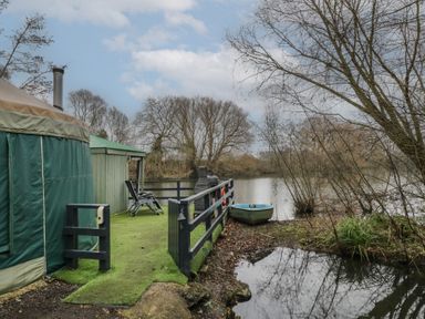 A green yurt and a deck area with chairs next to a small lake with a rowboat at The Lakeside Yurt in Beckford
