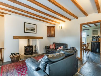 A living room with exposed wooden beams a wood stove two black leather sofas a red patterned rug and a view into a kitchen at Ty Capel Jeriwsalem Bronaber near Trawsfynydd