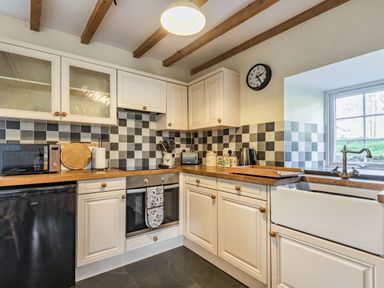 A kitchen with cream cabinets wooden countertops checkered tile backsplash a clock microwave and a window at Ty Capel Jeriwsalem Bronaber near Trawsfynydd